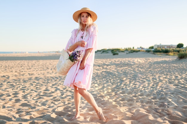 Imagen de altura completa de una chica rubia en un lindo vestido rosa bailando y teniendo fu en la playa. Sosteniendo flores y bolsa de paja.