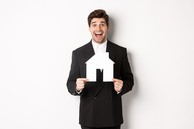 Foto gratuita imagen del apuesto agente inmobiliario en traje negro que muestra el mercado de casa, y mirando asombrado, vendiendo casas, de pie contra el fondo blanco.