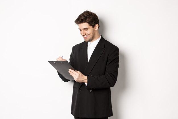 Imagen de apuesto hombre de negocios en traje firmando documentos, mirando el portapapeles y sonriendo, de pie contra el fondo blanco.