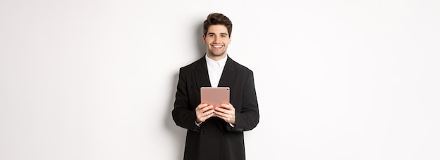 Imagen de un apuesto hombre de negocios con traje de moda sosteniendo una tableta digital y sonriendo de pie contra el whi