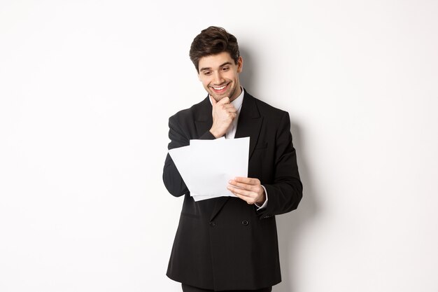 Imagen de un apuesto hombre de negocios en traje negro, mirando complacido con los documentos, leyendo el informe y sonriendo, de pie contra el fondo blanco.