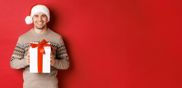 Imagen de un apuesto hombre sonriente con gorro de Papá Noel y suéter de invierno sosteniendo un regalo de Navidad y deseando felices fiestas de pie sobre un fondo rojo