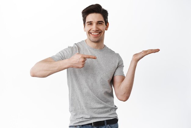 La imagen de un apuesto hombre sonriente señalando su mano vacía sosteniendo un artículo en la palma muestra su producto demostrando el espacio de la copia de pie satisfecho contra el fondo blanco