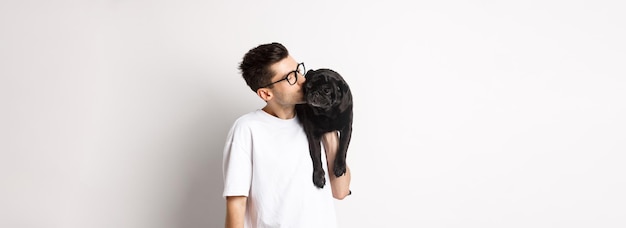 Foto gratuita imagen de un apuesto joven besando a su lindo pug negro sosteniendo al perro en el hombro parado sobre blanco b