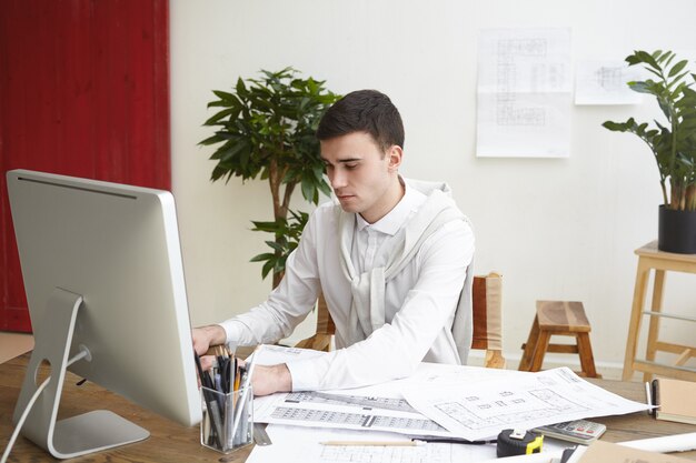 Imagen de un arquitecto masculino concentrado sentado en el escritorio con documentación y dibujos del proyecto, trabajando en un sistema CAD utilizando una computadora genérica. Concepto de personas, trabajo, ocupación, carrera y tecnología