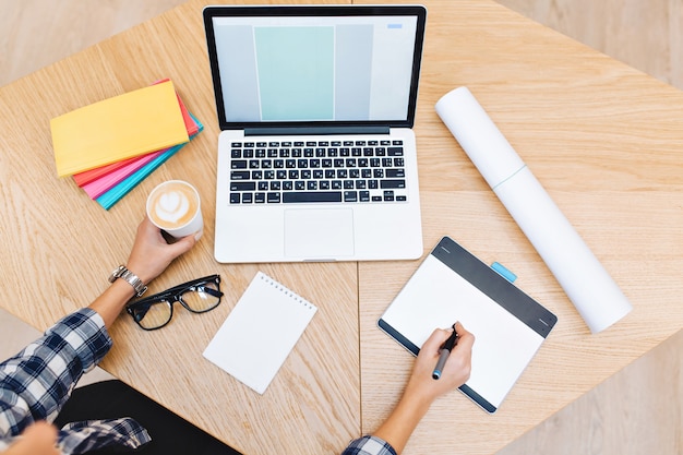 Imagen de arriba de cosas de trabajo en la mesa. Manos de mujer joven que trabaja con la computadora portátil, sosteniendo una taza de café. Cuadernos, gafas negras, trabajo duro, éxito, diseño gráfico.