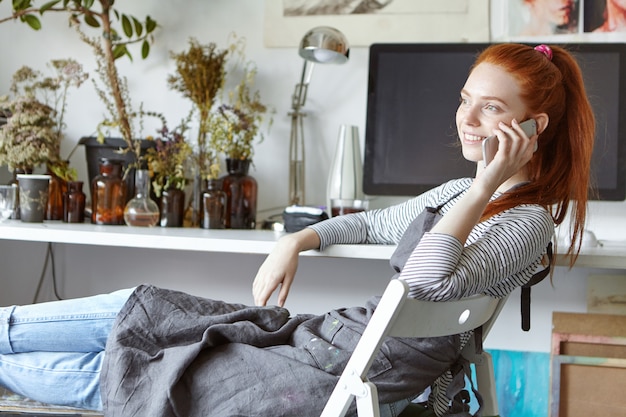 Foto gratuita imagen del artista europeo joven despreocupado confidente que se relaja en silla en estudio moderno, sonriendo alegremente mientras que teniendo una conversación telefónica agradable con un amigo. ocupación y tecnologías.