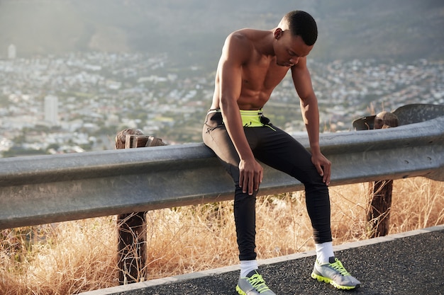 Imagen de un atleta de piel oscura cansado que usa mallas deportivas y zapatillas de deporte, tiene entrenamientos al aire libre, recupera el aliento después de correr, descansa en la señal de tráfico, mantiene las manos en las rodillas enfocadas hacia abajo. Estilo de vida saludable