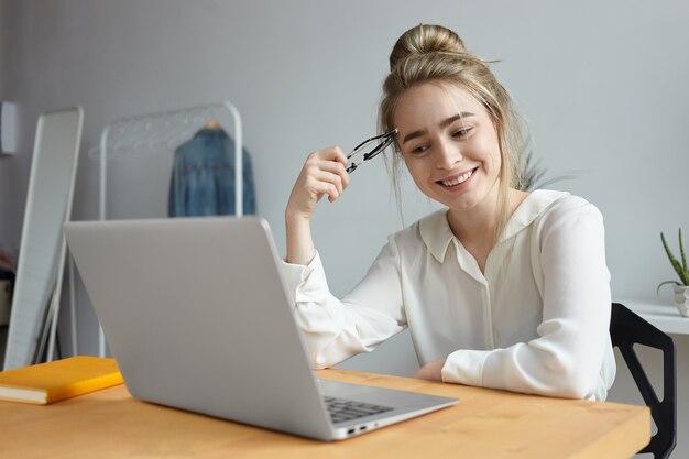 Imagen de una atractiva estudiante alegre con nudo de pelo navegando por Internet en una computadora portátil genérica, sentada en la mesa en casa, sonriendo ampliamente, viendo un blog, comunicándose con amigos en línea