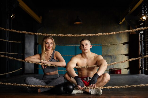 Imagen de la atractiva joven pareja atlética hombre y mujer sentados con las piernas cruzadas en el suelo dentro del ring de boxeo después de un entrenamiento intensivo, con una apariencia feliz y segura, con ropa deportiva elegante