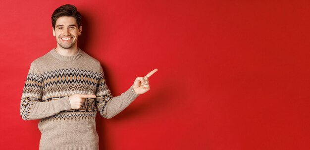 Imagen de un atractivo hombre sonriente, vestido con un suéter navideño, señalando con el dedo hacia la derecha y mostrando un anuncio de año nuevo, de pie sobre un fondo rojo.