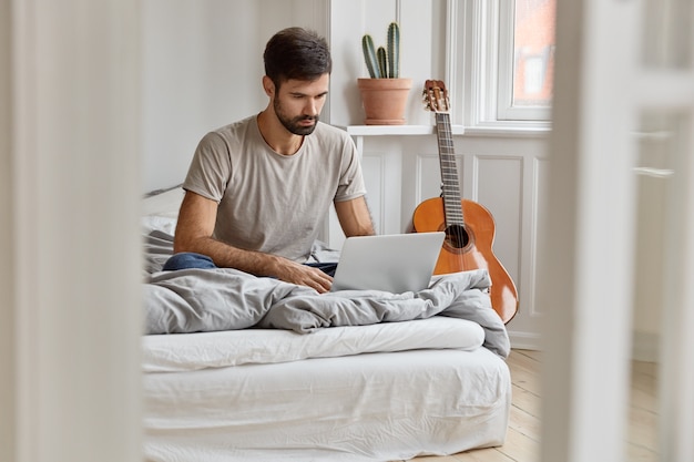 Imagen de un atractivo joven que se sienta en una cama cómoda, sincroniza archivos multimedia en una computadora portátil, trabaja como autónomo desde casa, disfruta del tiempo libre viendo películas, descansa en un apartamento acogedor.