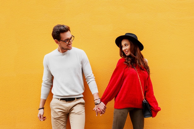 Foto gratuita imagen brillante de amantes posando en la pared amarilla. look de moda. estado de ánimo romántico. tomados de la mano. mujer joven con sincera sonrisa coqueteando con su novio. bolso de lujo.