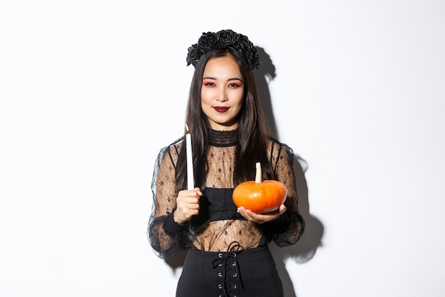 Imagen de bruja asiática sonriente astuta en traje gótico, sosteniendo una vela con calabaza y mirando a la cámara astucia, de pie sobre fondo blanco.