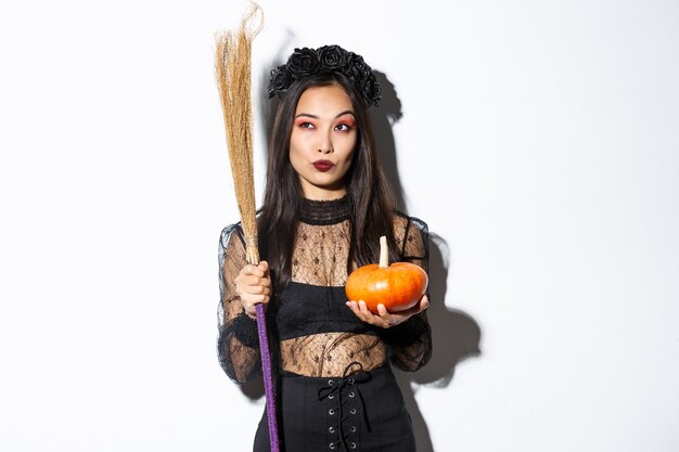 Imagen de bruja descarada en vestido de encaje gótico, sosteniendo escoba y calabaza, mirando en la esquina superior izquierda con banner de halloween, fondo blanco.