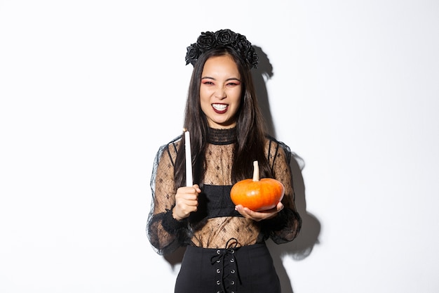 Imagen de la bruja malvada asiática en vestido de encaje gótico y corona negra, riendo y haciendo muecas, sosteniendo una vela con calabaza, celebrando halloween