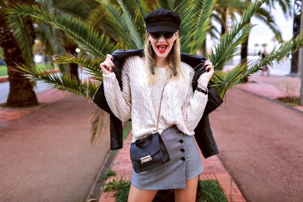 Imagen de la calle de moda de modelo con piernas largas con traje elegante glamour, tiempo de primavera, humor de viaje, palmeras alrededor, zapatillas de deporte, gorra, chaqueta de cuero, minifalda, suéter, accesorios y joyas.