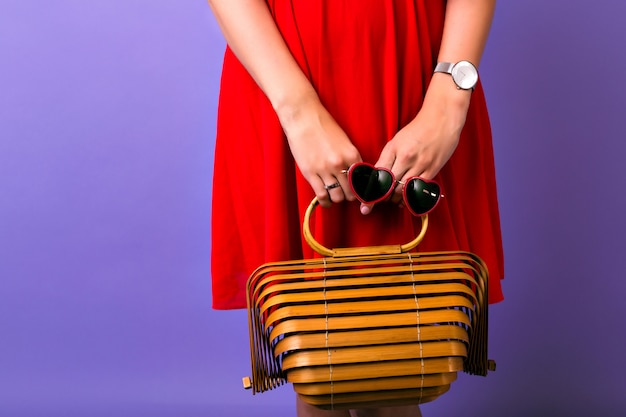 Imagen de cerca de moda o mujer con elegante vestido rojo brillante, sosteniendo un bolso de madera de paja y gafas de sol de corazón, reloj simple, fondo púrpura.