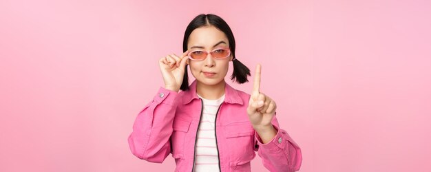 Imagen de una chica asiática con cara seria que muestra prohibir el gesto de regañar agitando el dedo apuntando hacia arriba expresa desaprobación de pie sobre fondo rosa