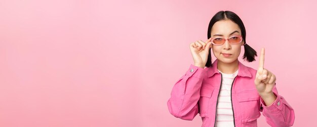 Imagen de una chica asiática con cara seria que muestra prohibir el gesto de regañar agitando el dedo apuntando hacia arriba expresa desaprobación de pie sobre fondo rosa