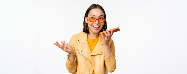 Imagen de una chica asiática feliz hablando en una grabación de altavoz traduciendo su voz con una aplicación de teléfono móvil hablando en un teléfono inteligente de pie dinámico sobre fondo blanco