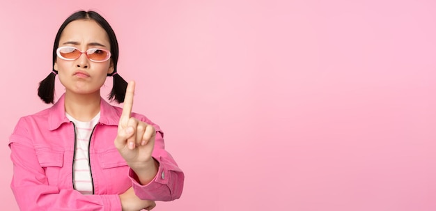 Imagen de una chica asiática seria y elegante con gafas de sol que muestra dejar de prohibir el gesto tabú que dice que no se para sobre fondo rosa
