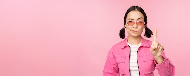 Imagen de una chica asiática seria y elegante con gafas de sol que muestra dejar de prohibir el gesto tabú que dice que no se para sobre fondo rosa