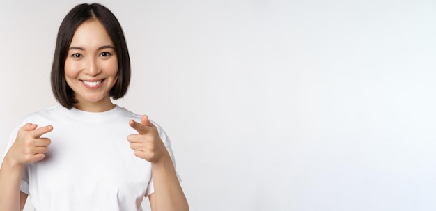 Imagen de una chica asiática sonriente señalando con el dedo a la cámara eligiendo invitarte a felicitarte de pie en camiseta sobre fondo blanco