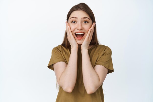 Imagen de una chica emocionada y sorprendida sonriendo, cogida de la mano en la cara emocionada, de pie en camiseta contra fondo blanco