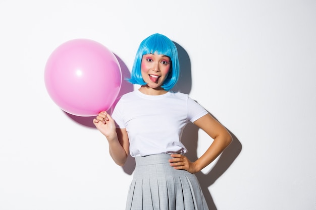 Imagen de una chica fiestera tonta con peluca azul celebrando la fiesta, sosteniendo un globo rosa y mostrando la lengua, fondo de pie.