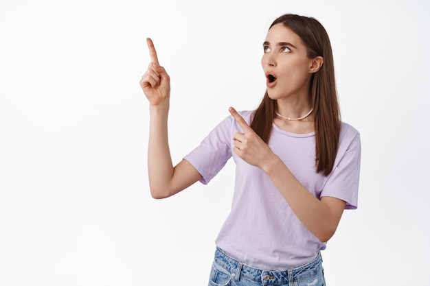 Imagen de una chica glam impresionada señalando y mirando el logotipo de la esquina superior izquierda, mostrando una pancarta y jadeando diciendo wow, asombrada con nuevos descuentos, publicidad, de pie sobre fondo blanco.