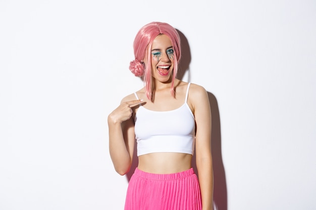 Imagen de una chica guapa descarada con peluca rosa y traje de fiesta, guiñando un ojo y apuntando a sí misma