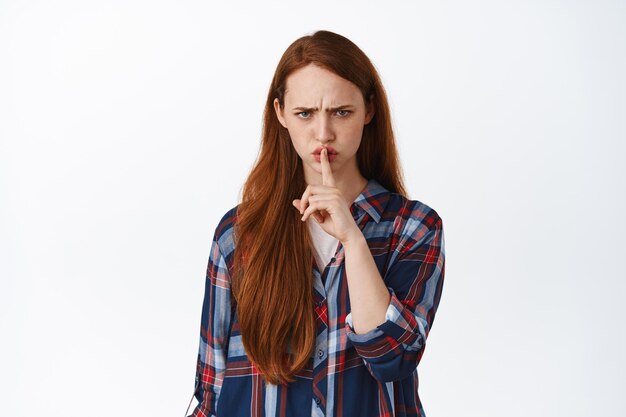 Imagen de una chica pelirroja enojada que se calla, dice shh y frunce el ceño disgustada, prohíbe, dice que se calle, se calla, no diga nada, de pie sobre fondo blanco