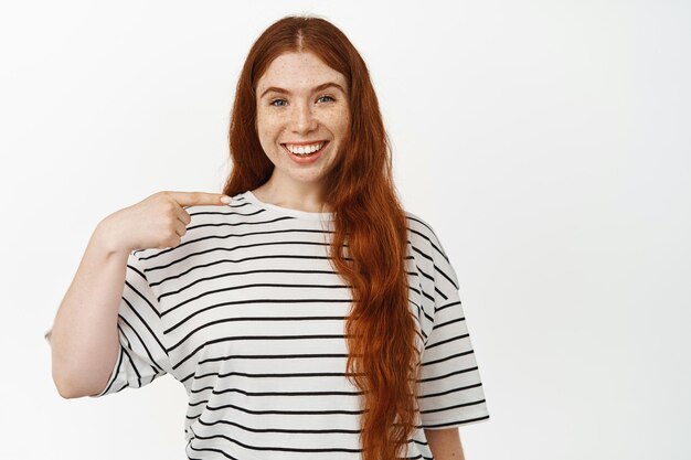 Imagen de una chica pelirroja sonriente con cabello largo y natural, apuntándose a sí misma con una expresión facial complacida, dientes blancos, mostrando algo en ella, de pie contra un fondo blanco.