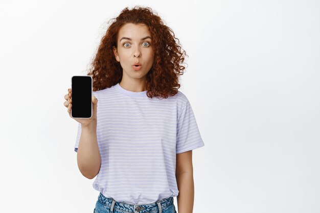 La imagen de una chica pelirroja sorprendida muestra un grito ahogado en la pantalla del teléfono vacío que muestra un anuncio en un teléfono inteligente de pie en una camiseta sobre fondo blanco