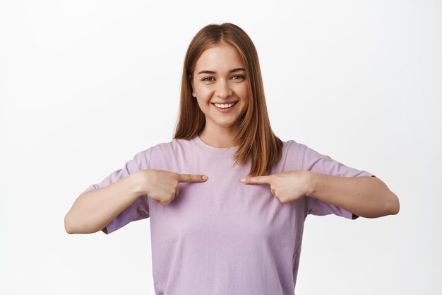 Imagen de una chica rubia con una camiseta apuntando al pecho, mostrándose a sí misma, con el logo en la ropa, sonriendo feliz, complacida contra un fondo blanco. copia espacio