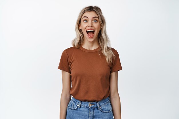 Imagen de una chica rubia que parece sorprendida y emocionada con la boca abierta y los ojos muy abiertos impresionados de pie en camiseta y jeans sobre fondo blanco
