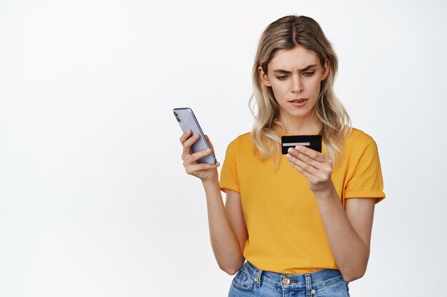 Imagen de una chica rubia seria sosteniendo un teléfono móvil mirando el número de la tarjeta de crédito con la cara concentrada tratando de pagar en línea de pie sobre un fondo blanco