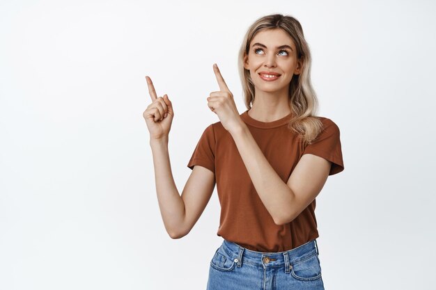 Imagen de una chica rubia sonriente señalando con el dedo y mirando la esquina superior izquierda con una expresión de cara satisfecha y pensativa de fondo blanco