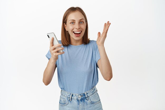 Imagen de una chica rubia sorprendida saltando de la felicidad, una gran noticia en un teléfono móvil sosteniendo un teléfono inteligente y regocijándose de haber ganado en línea de pie contra el fondo blanco