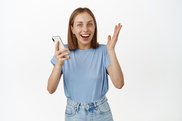 Imagen de una chica rubia sorprendida saltando de la felicidad, una gran noticia en un teléfono móvil sosteniendo un teléfono inteligente y regocijándose de haber ganado en línea de pie contra el fondo blanco