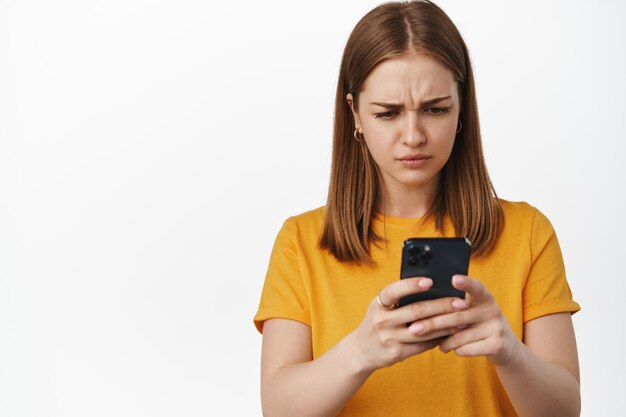 La imagen de una chica rubia se ve confundida con el teléfono móvil, notificación de mensaje extraño en el teléfono inteligente, problema en el teléfono celular, parada en una camiseta amarilla contra fondo blanco.