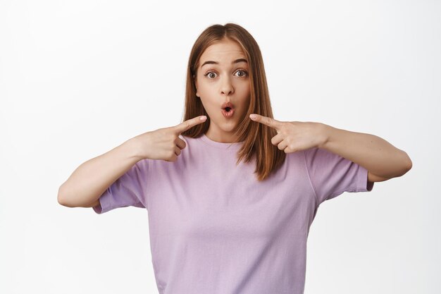 La imagen de una chica rubia se ve sorprendida, dice wow y señala con el dedo a la boca, parada en una camiseta de verano contra un fondo blanco. copia espacio