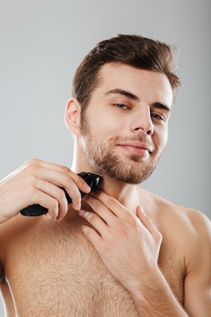 Imagen de un chico adulto guapo haciendo un procedimiento de higiene y salud al afeitarse las cerdas con una recortadora sobre una pared gris