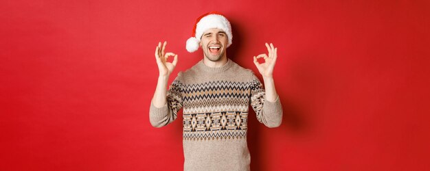 Imagen de un chico alegre y atractivo con suéter y gorro de Papá Noel deseando feliz navidad mostrando señales de estar bien