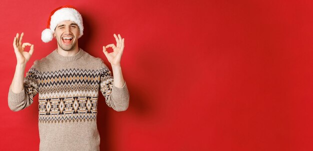Imagen de chico atractivo alegre en suéter y gorro de Papá Noel, deseando feliz Navidad, mostrando signos de bien y guiñando un ojo a la cámara, celebrando el año nuevo, de pie sobre fondo rojo