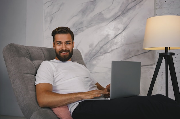 Imagen de un chico atractivo con barba espesa que disfruta de la comunicación en línea a través de las redes sociales, utilizando una conexión a Internet de alta velocidad en una computadora portátil, mirando a la cámara con una sonrisa de satisfacción y confianza