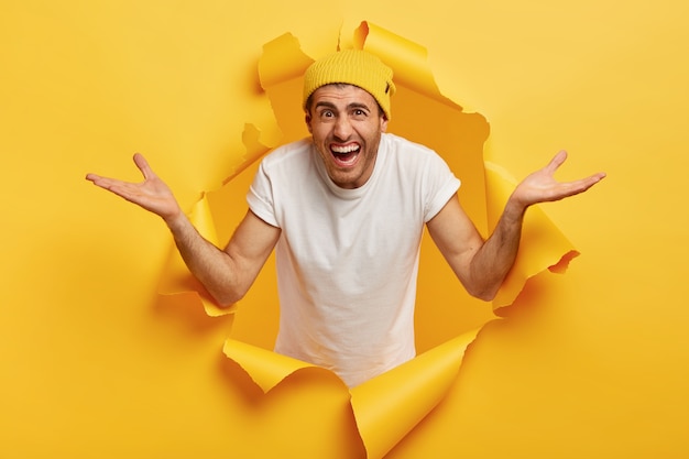 La imagen de un chico emocional usa sombrero y camiseta blanca informal, extiende las manos hacia los lados, tiene una expresión facial alegre y despistada