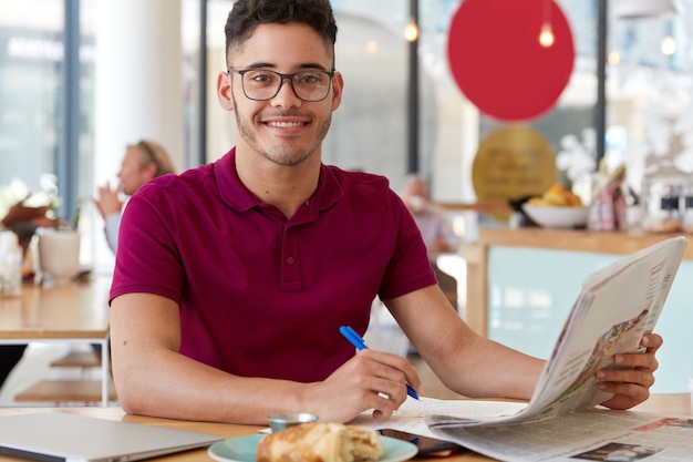 La imagen del chico guapo y complacido lee las últimas noticias en el periódico, graba algunas notas en el bloc de notas, usa gafas y camiseta, disfruta de un delicioso croissant. Personas y concepto de trabajo