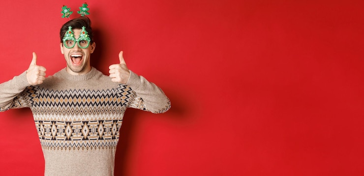 Foto gratuito imagen de un chico guapo emocionado con gafas de fiesta y suéter de navidad mostrando pulgares arriba alabando la gran celebración de año nuevo de pie sobre un fondo rojo